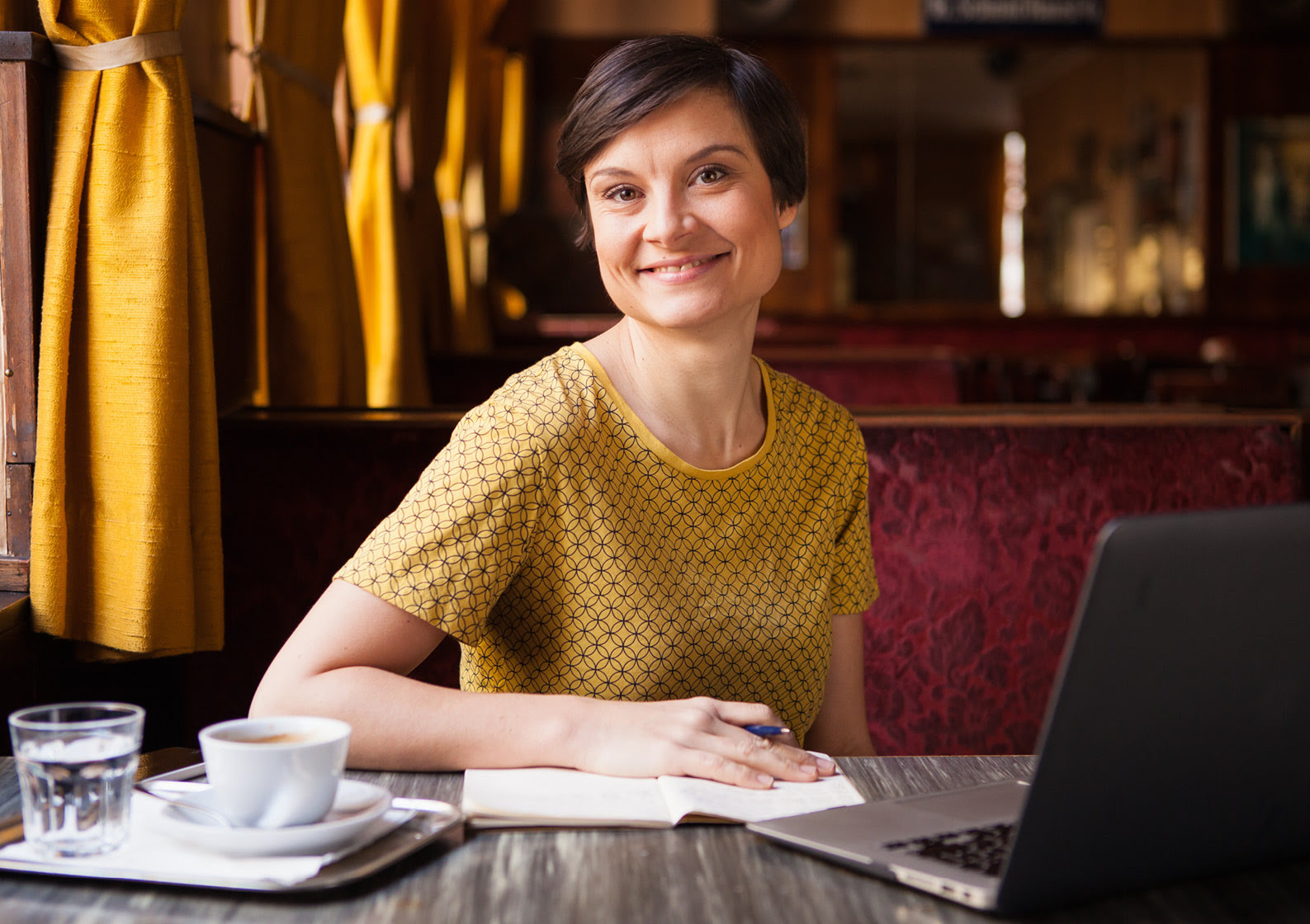 Lena Raubaum sitzt mit Kaffee, Wasserglas, Notizbuch und Laptop entspannt in einem elegant eingerichteten Caf&eacute; mit samtenen, roten B&auml;nken und senfgelben Vorh&auml;ngen.