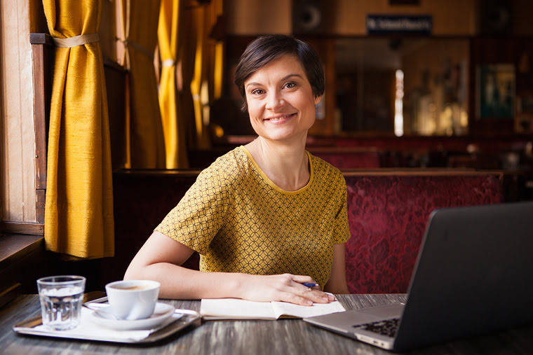 Portrait Lena Raubaum lächelnd in Café mit Laptop, Kaffee und Notizblock